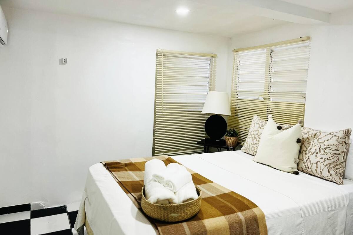 A hotel room with a bed, a bedside table with a lamp, and two windows with blinds.