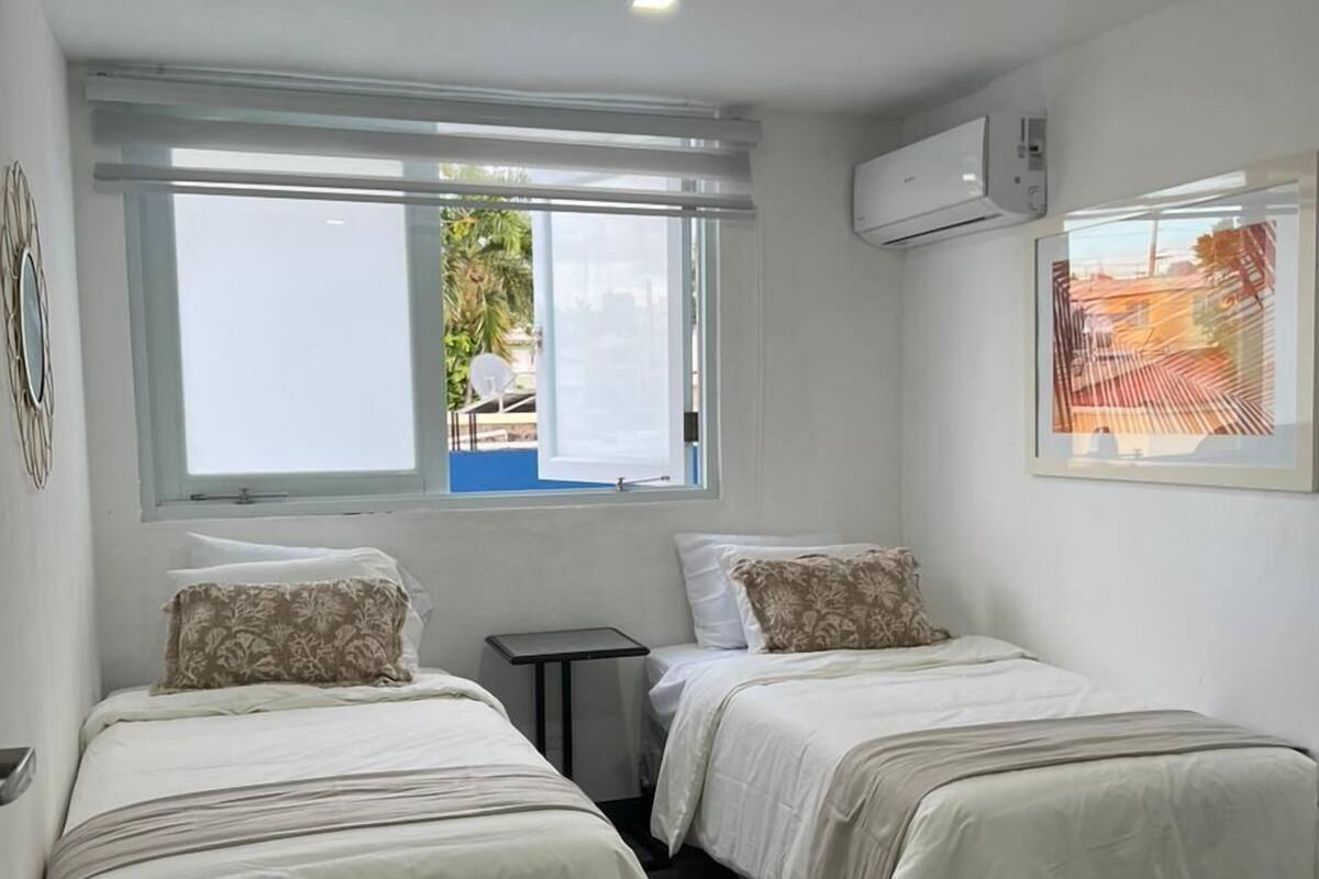 A hotel room with two beds, a small table, a window with a view, and an air conditioning unit.