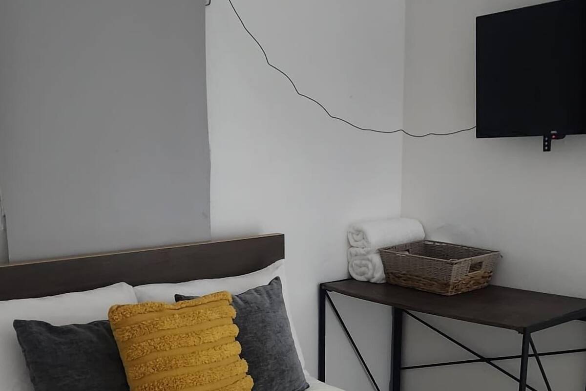 A neatly made bed with a white and beige striped blanket, two pillows (one yellow and one gray), and a wooden headboard. A small table with a basket and a rolled towel is beside the bed.