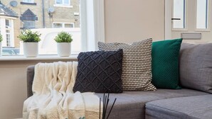 En soffa med en beige pläd, en mörk, mönstrad kudde, en grön kudde och en vit, mönstrad kudde. På fönsterbrädan står krukväxter.