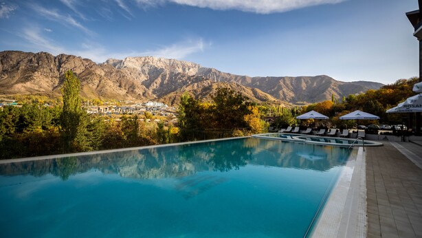Piscina con montañas al fondo, sillas de descanso y sombrillas.