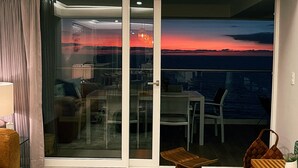 Een moderne woonkamer met een grote ronde poef, een houten bankje en een glazen deur die leidt naar een balkon met uitzicht op de ondergaande zon boven het water.