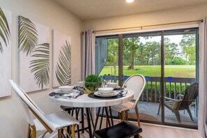 A dining area with a table set for two, a chair, a bench, and a view of a grassy outdoor area through large glass doors.