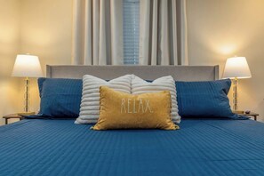 A bed with a blue bedspread, two pillows, and a 'RELAX' pillow. There are two bedside lamps and a window with blinds.