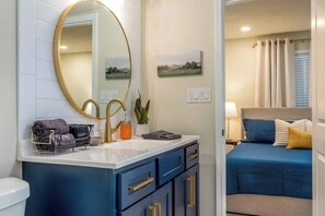 A bathroom with a large mirror, a sink, and a toilet. There is a bed with blue bedding and a yellow pillow in the adjacent room.