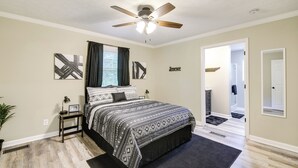 Un dormitorio con cama, ventilador de techo, ventana con cortinas y un pasillo que lleva a otra habitación.