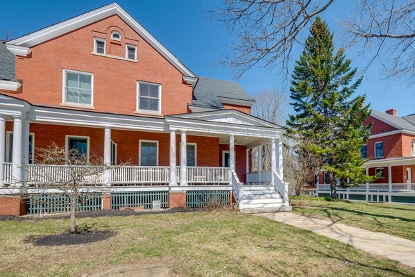 A two-story brick house with a front porch, a small tree in front, and a clear blue sky.