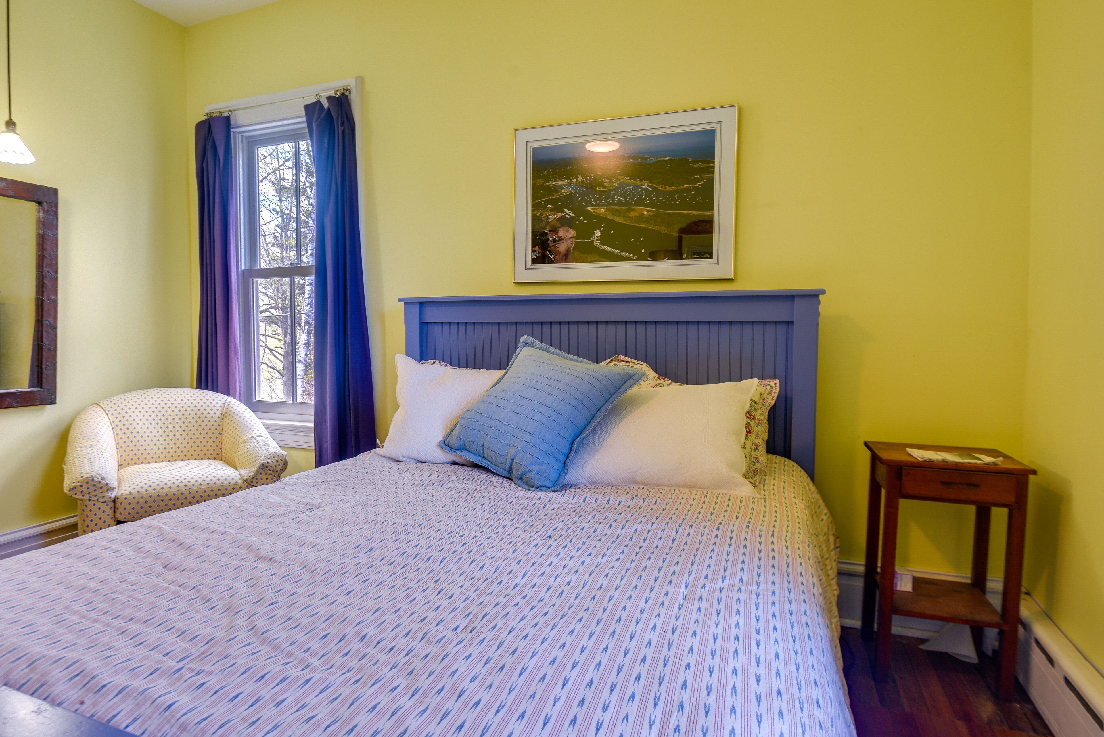 A bedroom with a bed, a chair, a nightstand, a window with curtains, and a framed picture on the wall.