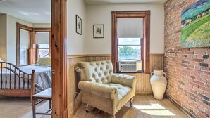 A room with a wooden chair, a cushioned armchair, a large vase, and a painting on a brick wall. - Historic Apartment - Half Mi to Lake Champlain! (Plattsburgh)