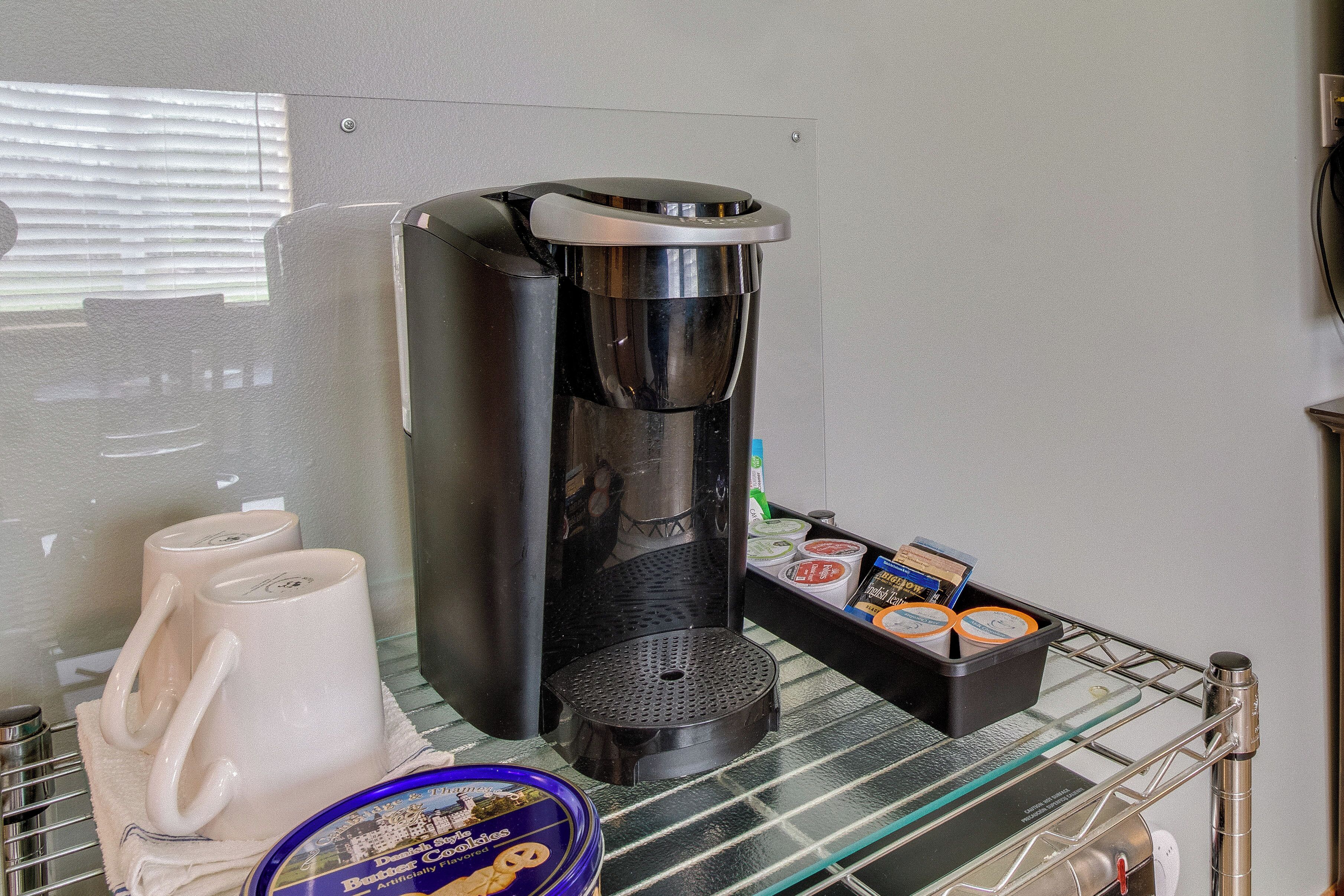A coffee machine with a tray of mugs and a container of cookies on a glass shelf.