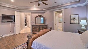 A bedroom with a bed, dresser, mirror, TV, ceiling fan, and a door leading to another room.