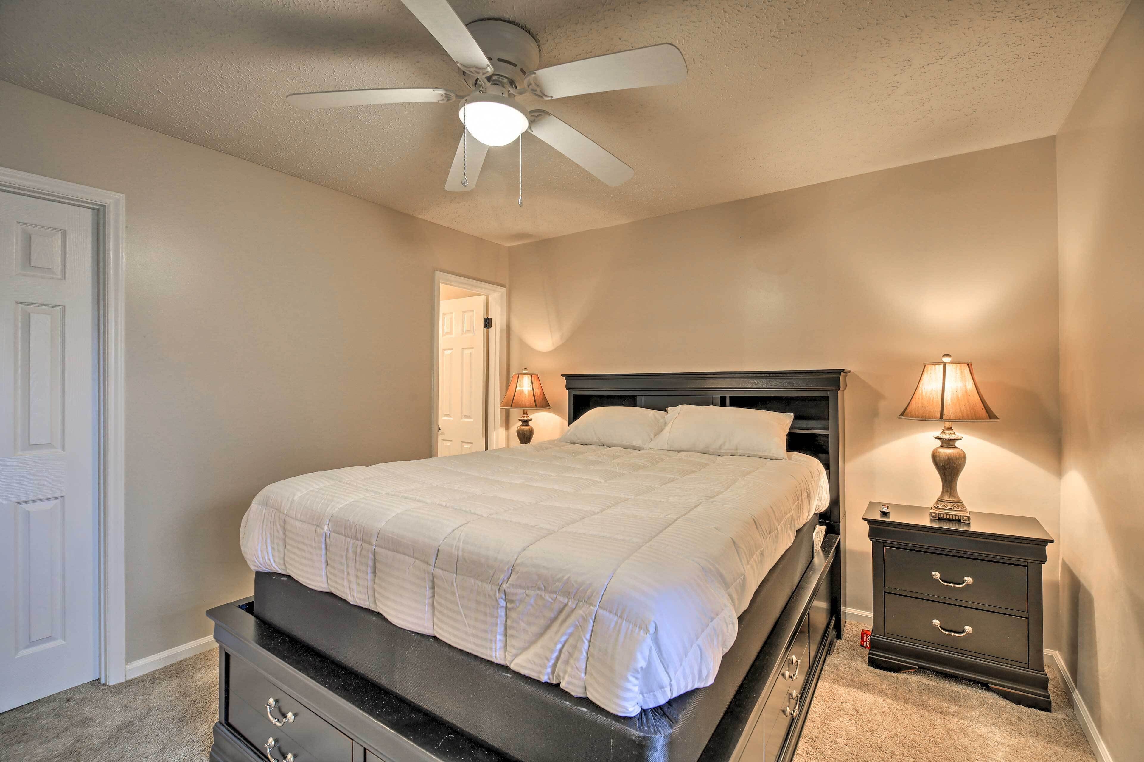 A bedroom with a bed, a nightstand with a lamp, a ceiling fan, and a door.