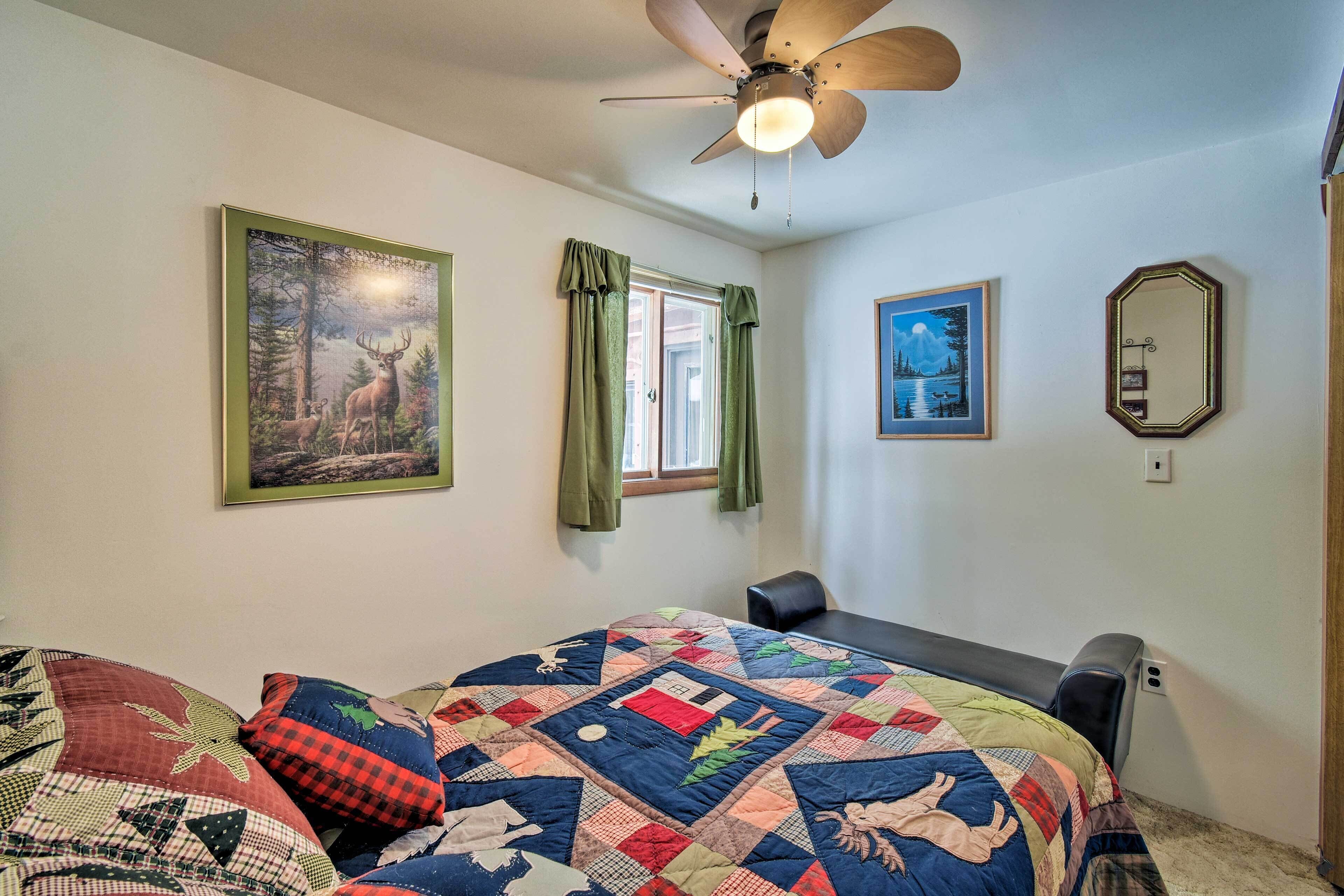 A bedroom with a large bed, a ceiling fan, a window with curtains, and two framed pictures on the wall.