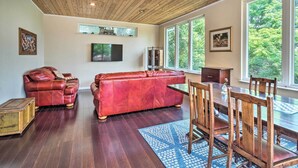 Ein geräumiges Wohnzimmer mit Holzbalkendecke, einem roten Ledersofa, einem Esstisch mit Holzstühlen und großen Fenstern mit Blick ins Freie.