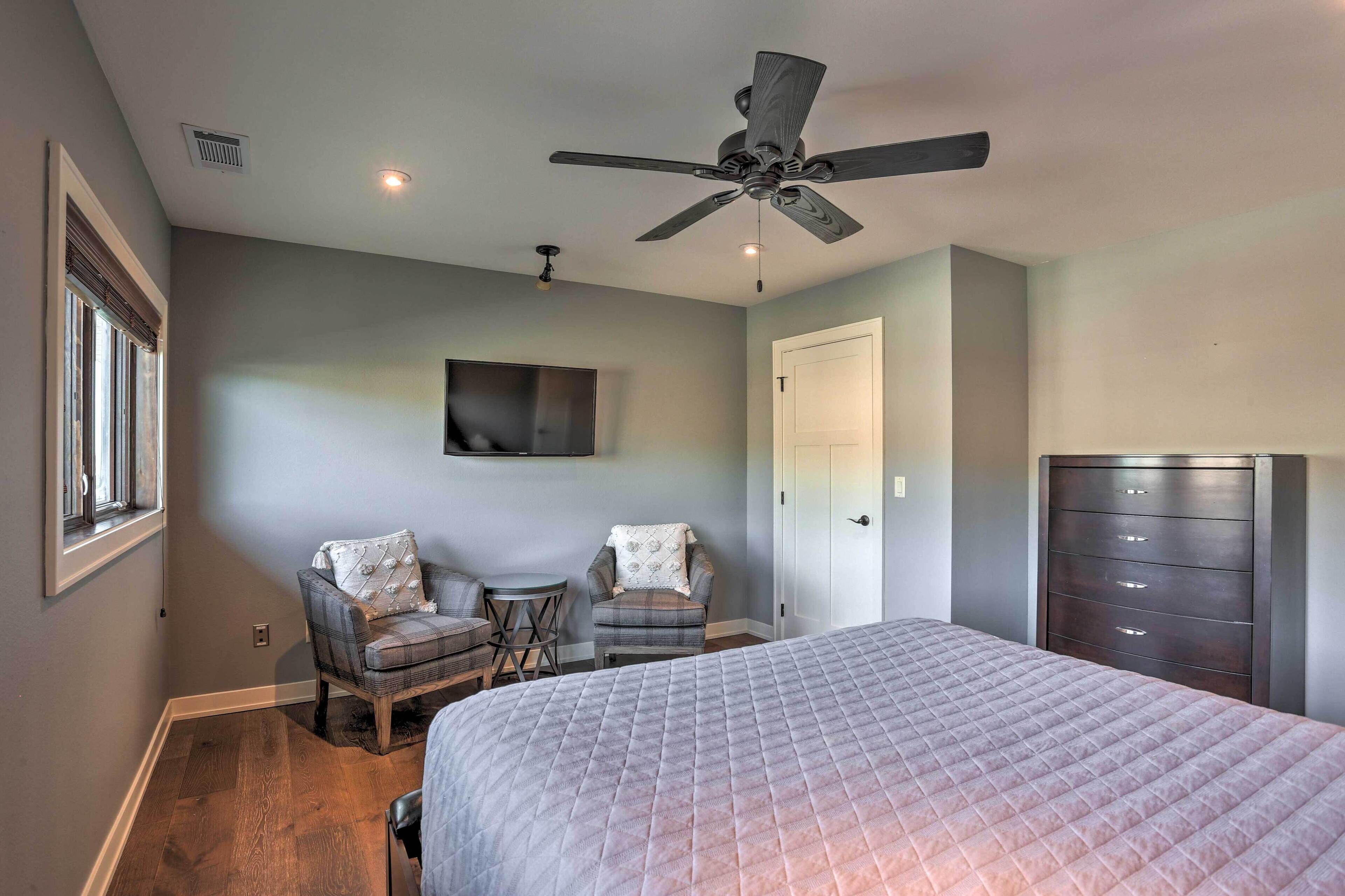 A bedroom with a bed, two chairs, a ceiling fan, a TV, and a dresser.