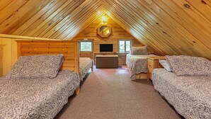 A cozy attic bedroom with wooden walls and a pitched roof. There are two beds with patterned bedding, a small sofa, and a television.