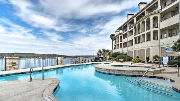 A swimming pool with a stone border, a palm tree, and a building with balconies in the background. - Lakefront Lago Vista Condo w/ Resort Amenities! (Leander)