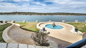 A landscaped area with a circular fountain, a paved walkway, and a view of a body of water and distant land. - Lakefront Lago Vista Condo w/ Resort Amenities! (Leander)