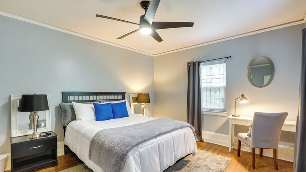 Quarto com cama, mesas de cabeceira, escrivaninha, cadeira, ventilador de teto e janela com cortinas.