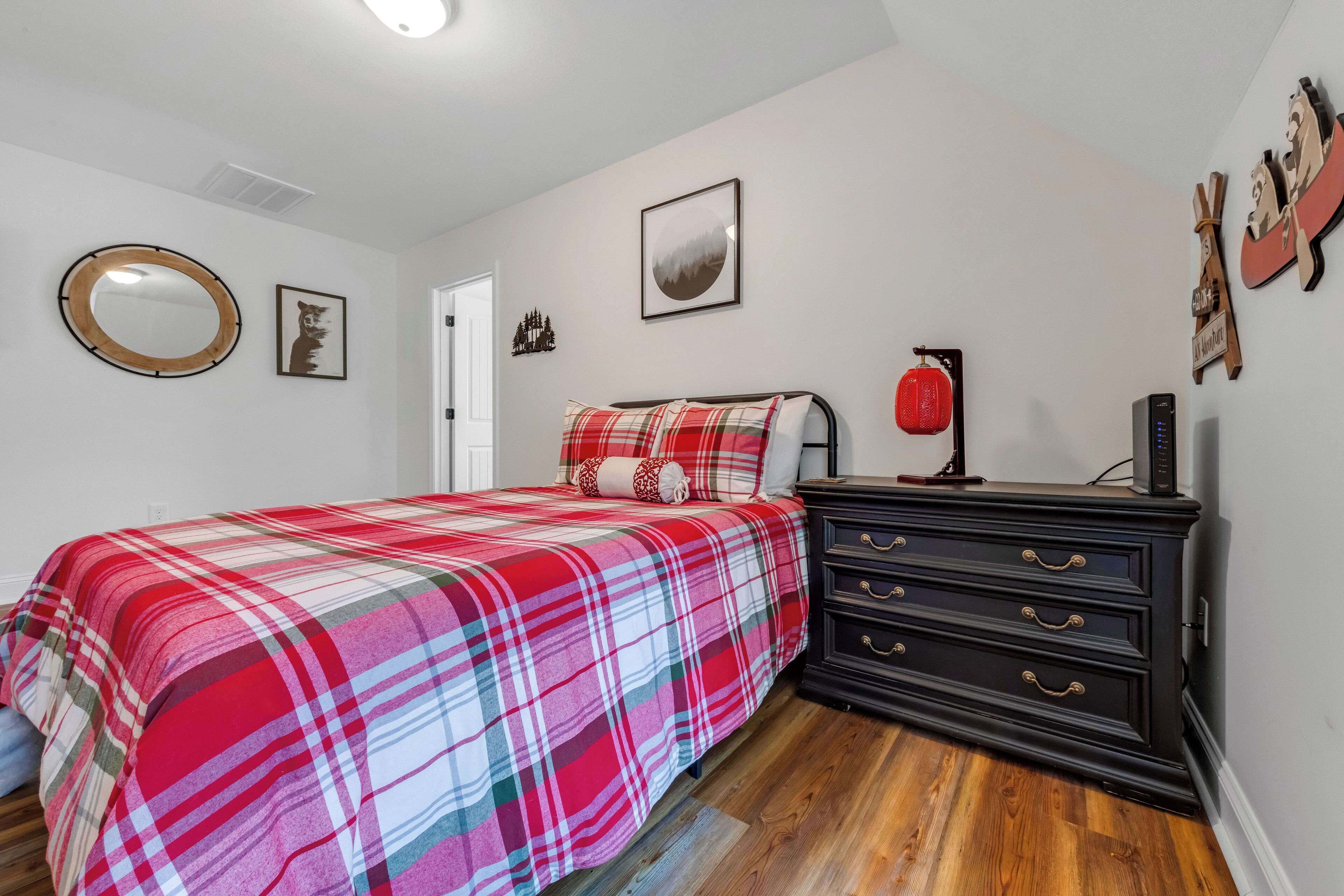 A bedroom with a plaid bedspread, a dresser, a mirror, and wall art.