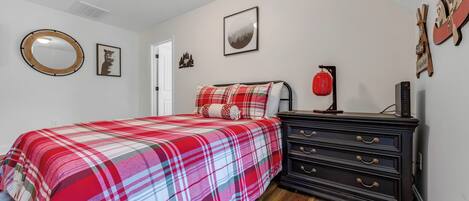 A bedroom with a plaid bedspread, a dresser, a mirror, and wall art.
