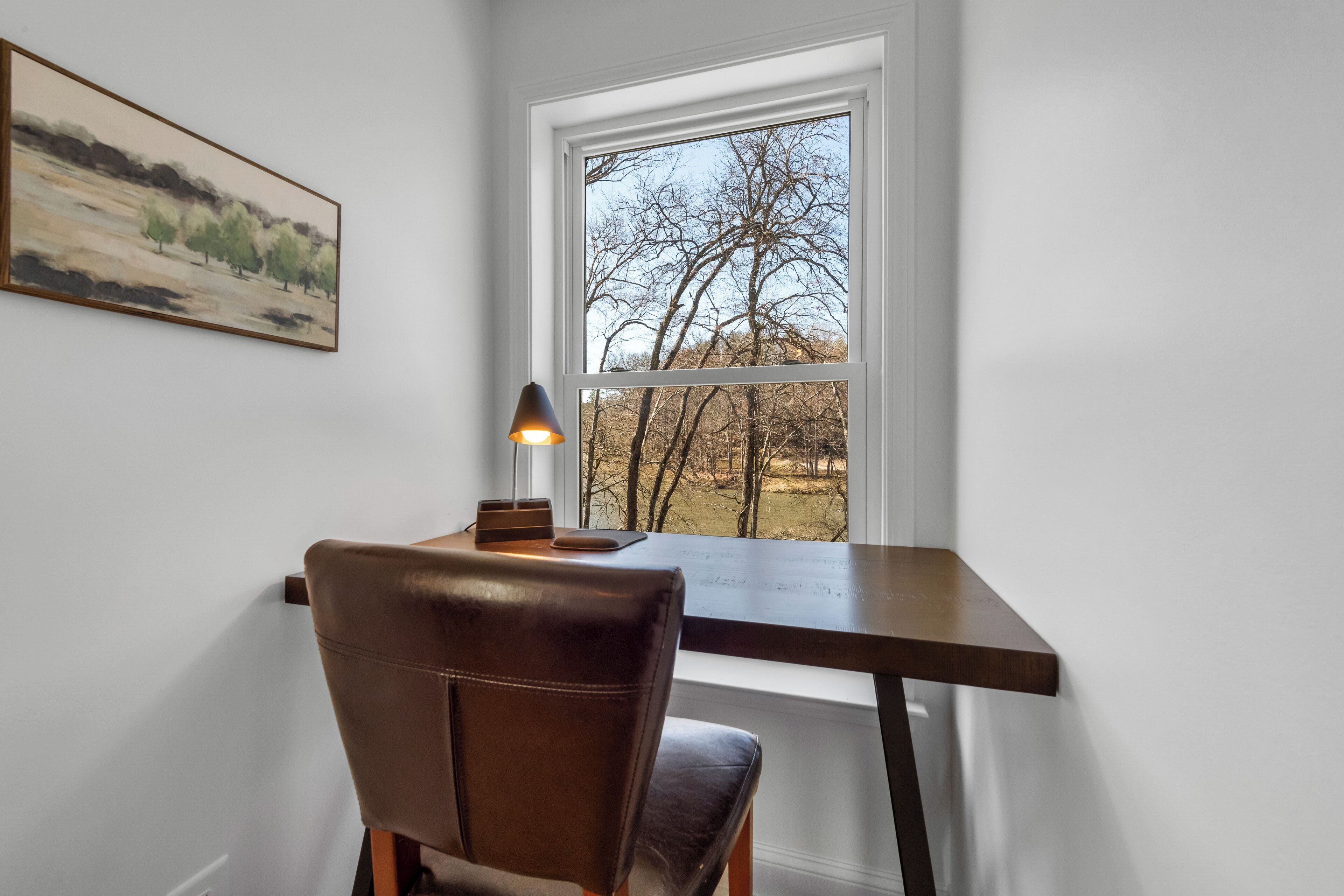 Une pièce comprenant un bureau, une chaise, une lampe et une fenêtre donnant sur des arbres.