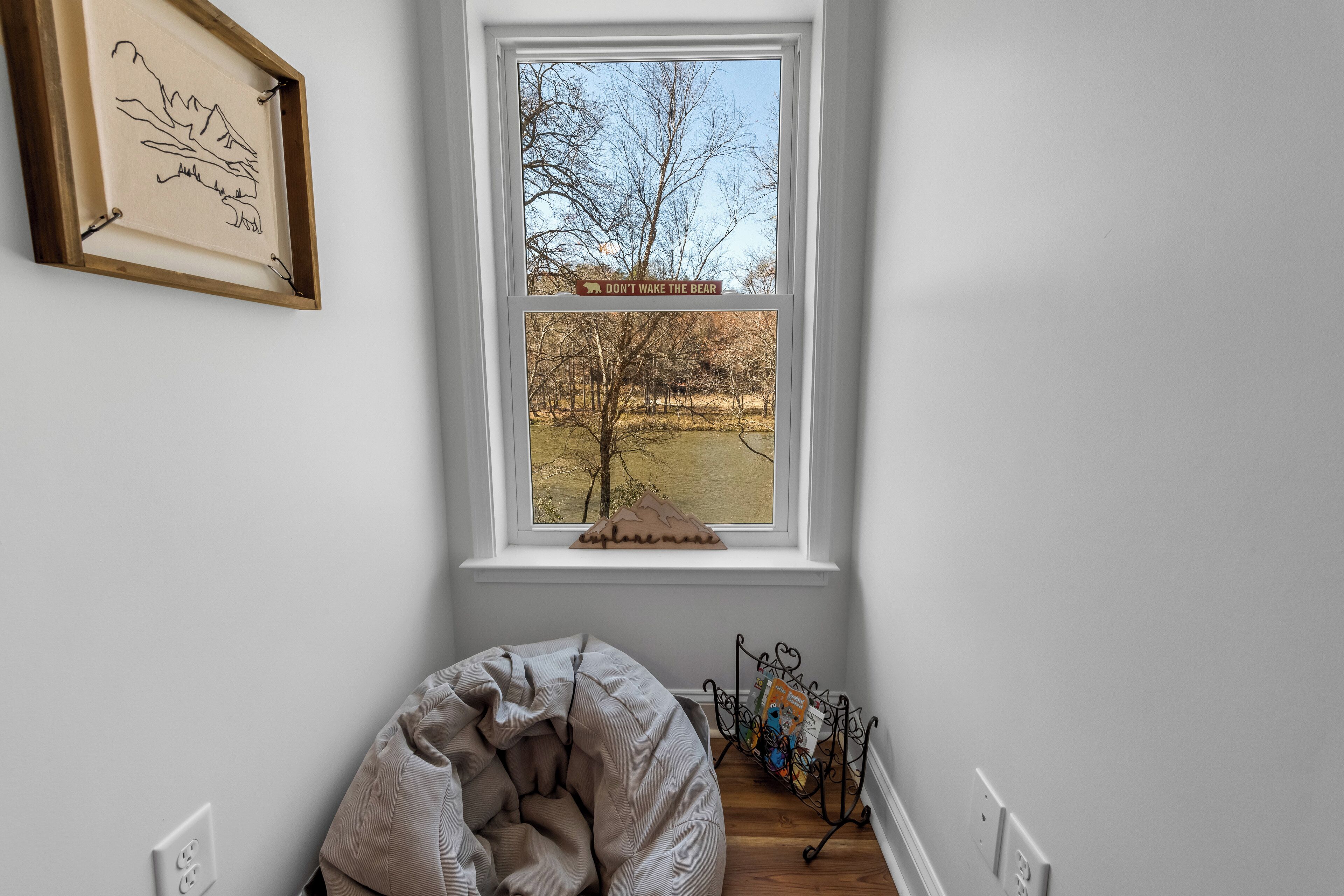 A room with a window overlooking a forest, a framed picture on the wall, and a bookshelf with books.