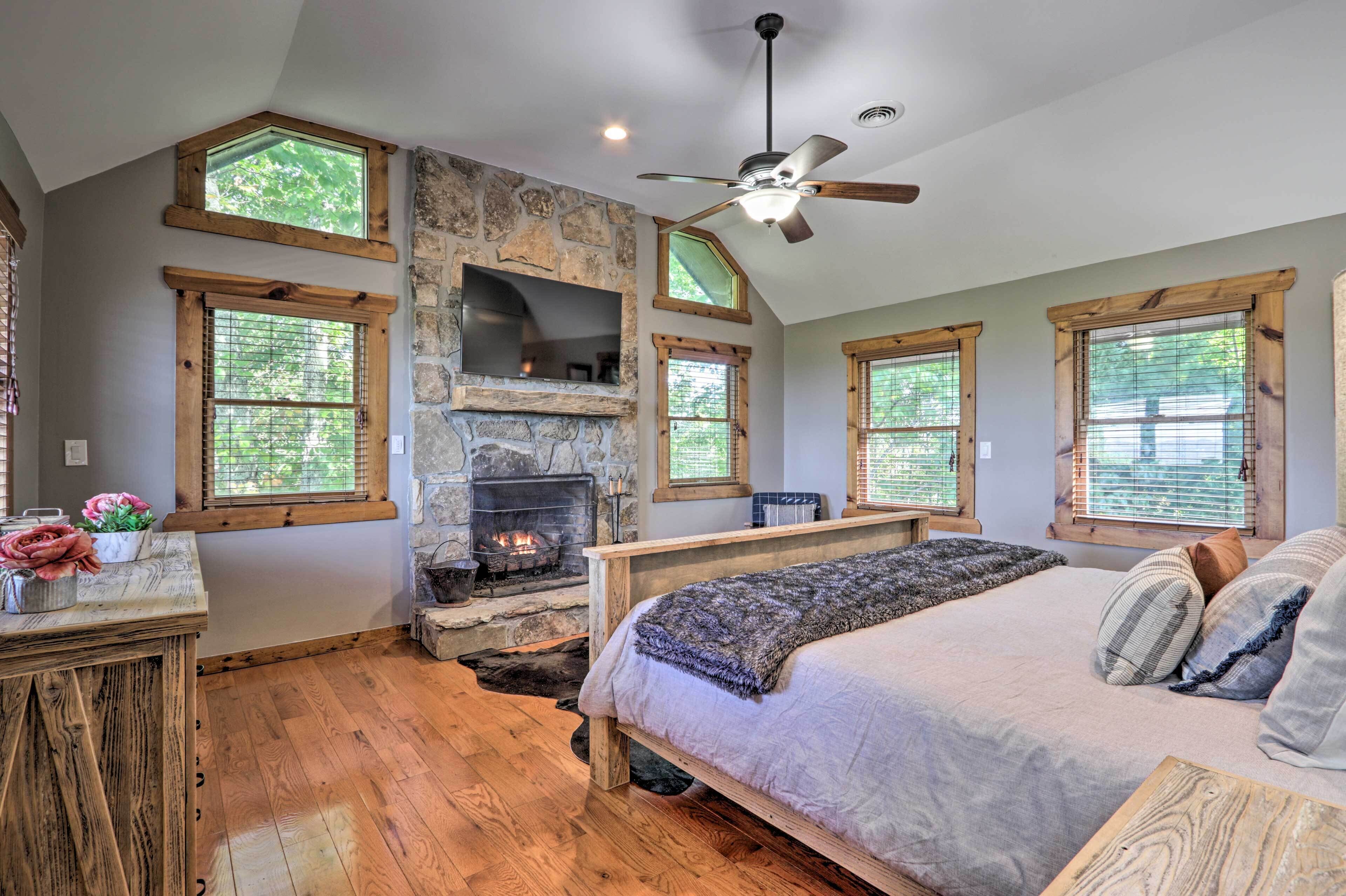 A bedroom with a stone fireplace, wooden beams, a bed, and large windows.