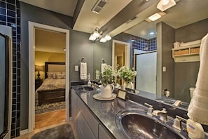A modern bathroom with a dark countertop, two sinks, a large mirror, and a bed visible through an open door.