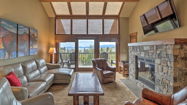 A spacious living room with a stone fireplace, leather sofas, a wooden coffee table, and a large window overlooking a scenic view. - Expansive Bigfork Resort Retreat on Flathead Lake! (Bigfork)