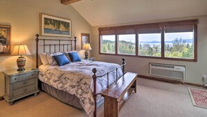 A bedroom with a large bed, a wooden bench, a nightstand, a window with a view, and a wall-mounted heater. - Expansive Bigfork Resort Retreat on Flathead Lake! (Bigfork)
