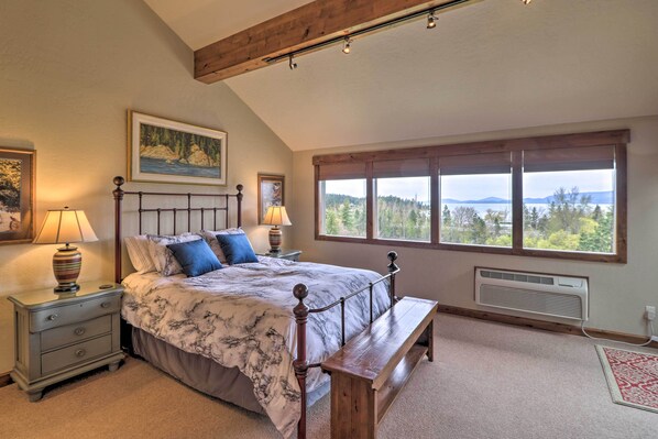 A bedroom with a large bed, a wooden bench, a nightstand, a window with a view, and a wall-mounted heater. - Expansive Bigfork Resort Retreat on Flathead Lake! (Bigfork)