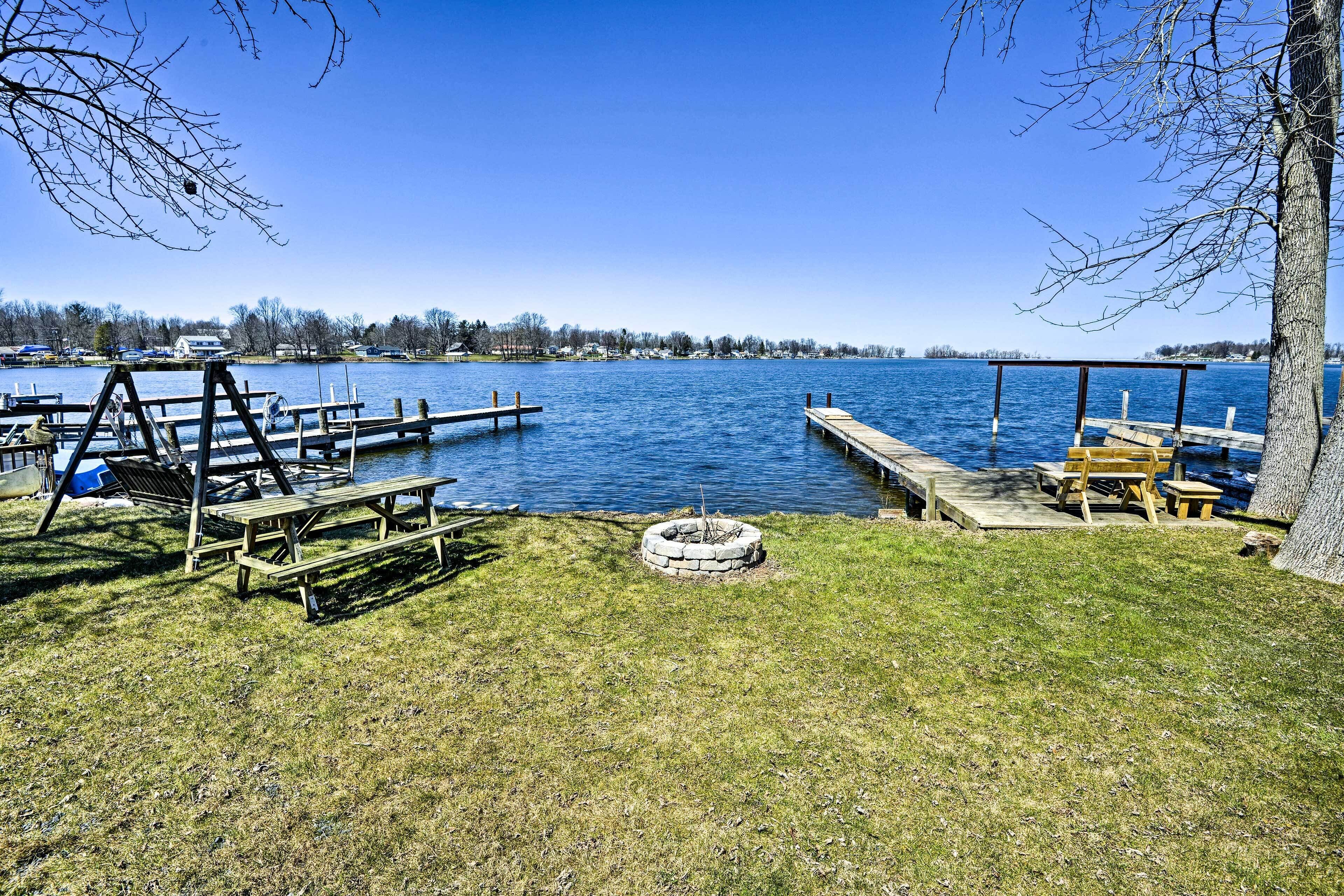 A lakeside picnic area with wooden benches, a fire pit, and a dock extending into the water.