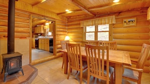 A log cabin interior with a dining area, wooden chairs, a table, a stove, and a kitchen visible through an open door. - 'honey's Cabin' in Mcleod: Fish, Hike & Views! (McLeod)