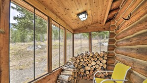 A log cabin interior with large windows, a chair, and a stack of firewood. - 'honey's Cabin' in Mcleod: Fish, Hike & Views! (McLeod)