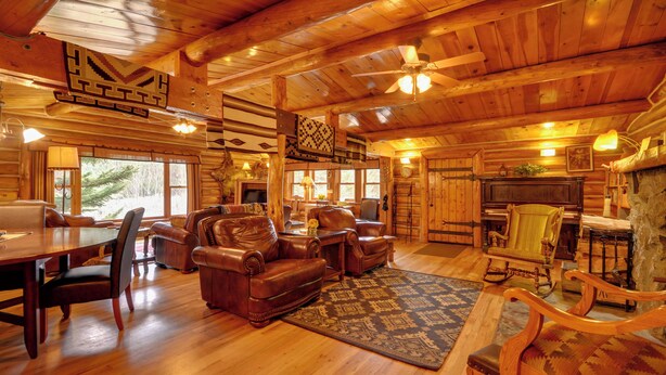 A log cabin interior with a dining area, leather sofas, a fireplace, and a large window offering a view of trees. - 'honey's Cabin' in Mcleod: Fish, Hike & Views! (McLeod)