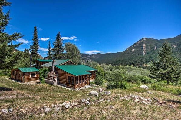 A log cabin with a green roof, surrounded by a forested landscape and mountains in the background.
