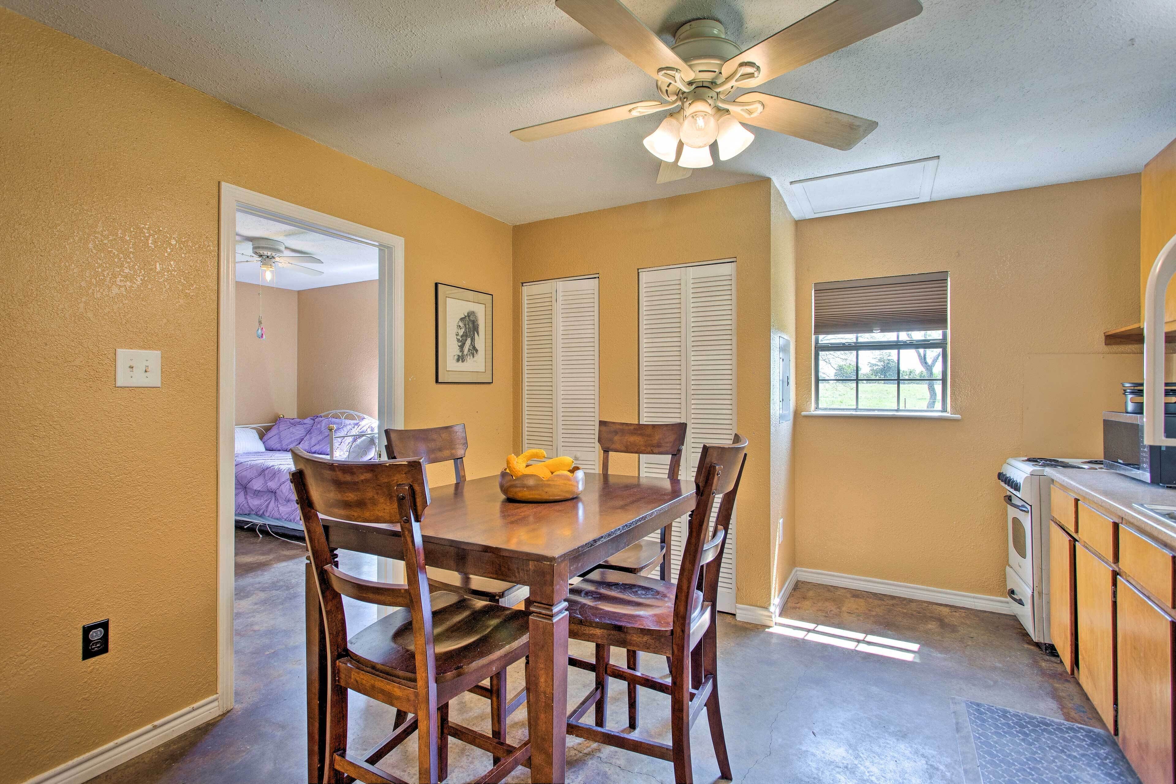 A dining area with a wooden table and chairs, a ceiling fan, and a kitchen with a microwave and oven.