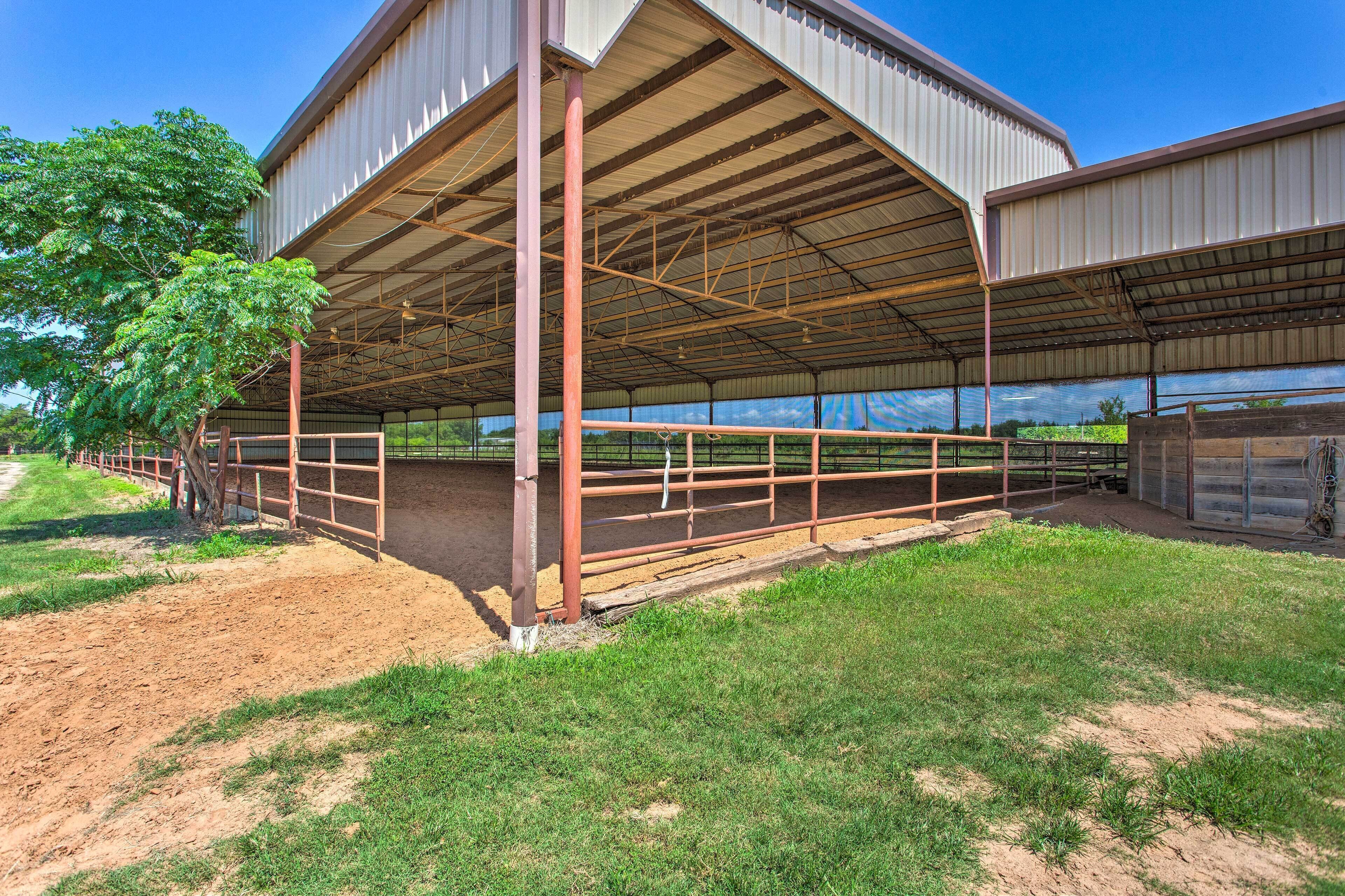 A stable with a metal roof and open sides, surrounded by a fenced area with a dirt path leading to it.