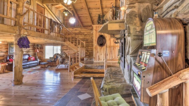 A rustic lodge interior with wooden floors, a stone fireplace, a wooden staircase, and a jukebox. - Secluded Belle Fourche Cabin: Group Friendly! (Belle Fourche)