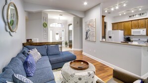 A modern living room with a blue sectional sofa, a round wooden tray table, and a kitchen area with wooden cabinets and a white refrigerator. - Ocean Springs Home w/ Fire Pit & Game Room! (Biloxi)