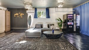 A modern hotel room with a sofa, a coffee table, a decorative wall piece, a window with curtains, and a vending machine.