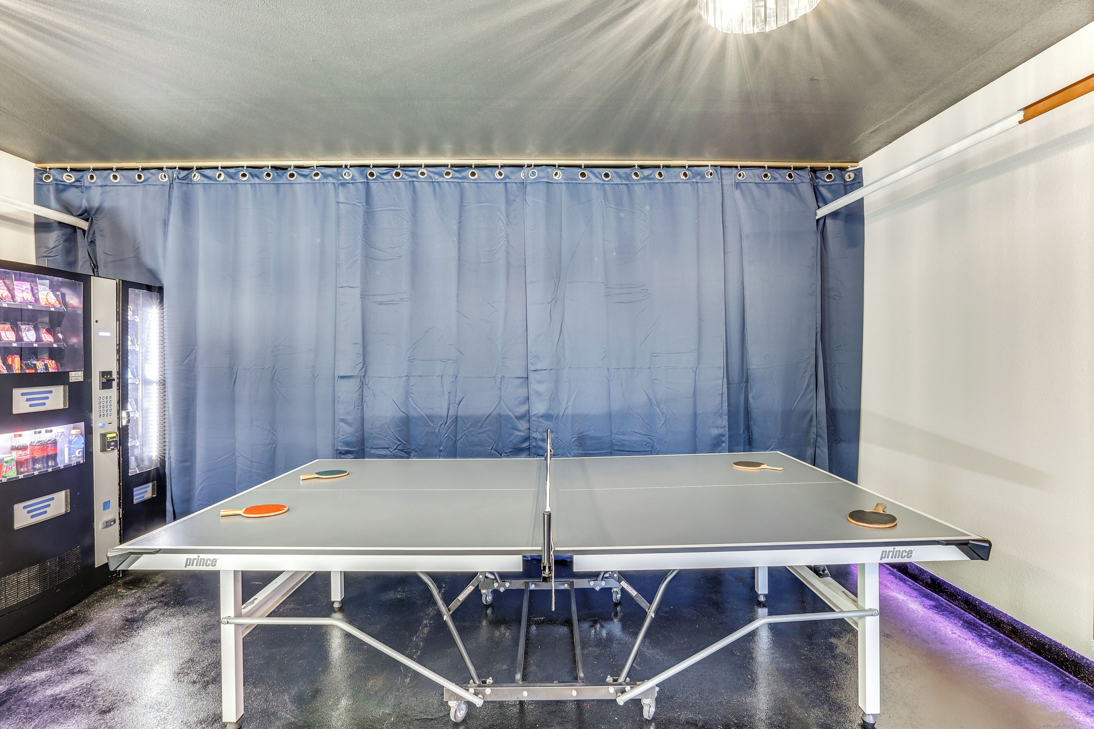 Une table de ping-pong avec deux raquettes, un distributeur automatique et un rideau.