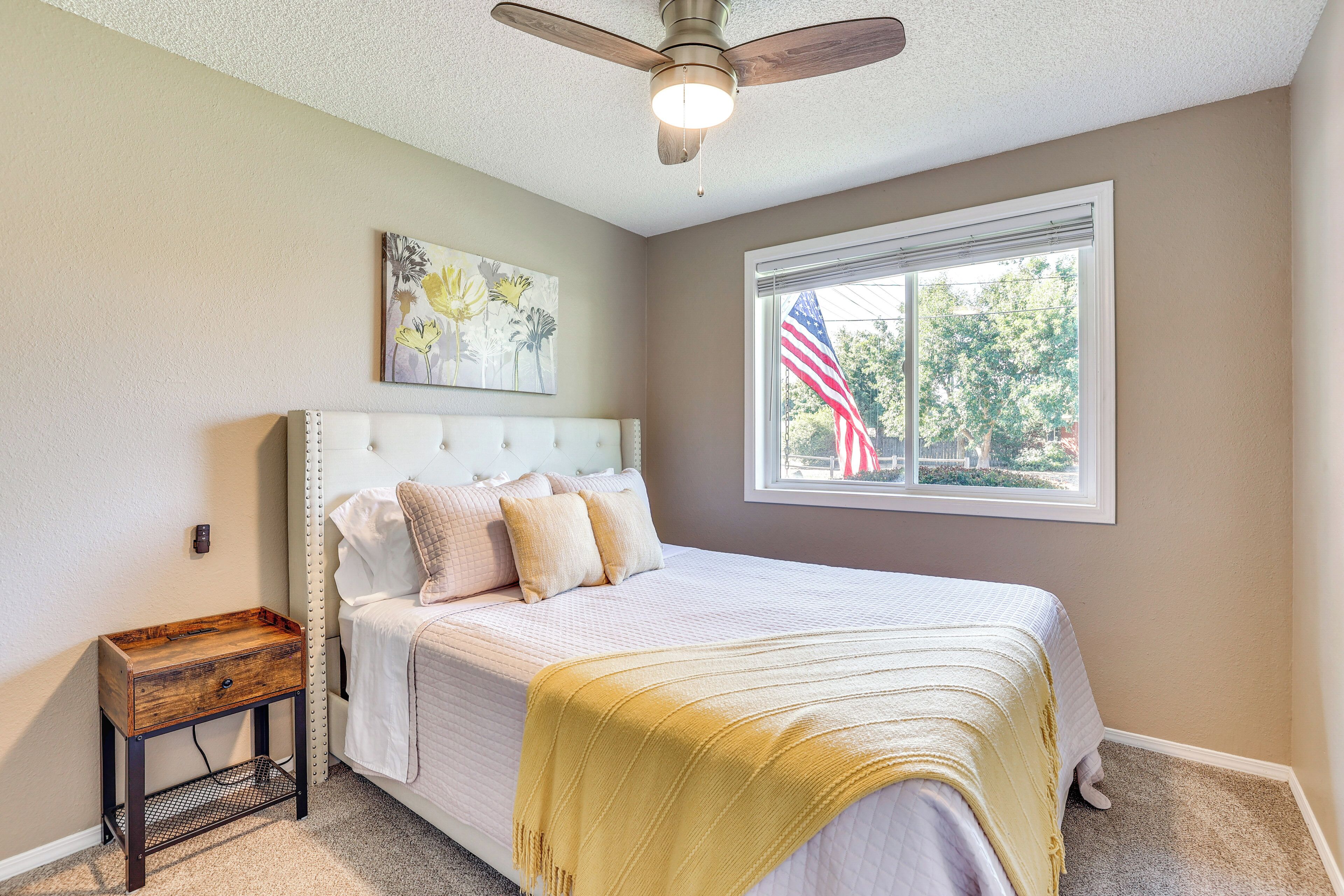 A bedroom with a bed, a nightstand, a ceiling fan, a window with an American flag, and a painting on the wall.