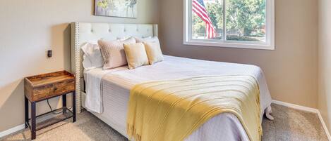 A bedroom with a bed, a nightstand, a ceiling fan, a window with an American flag, and a painting on the wall.