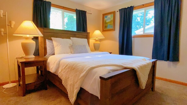 A neatly made bed with white linens, two bedside lamps, a wooden nightstand, and a painting on the wall. - Fantastic Oasis Home in Stevens Point with fully enclosed fence for backyard! (Stevens Point)