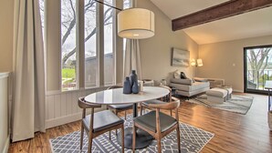 A modern living room with a round dining table, wooden chairs, a sofa, and a large window with a view of the outdoors. - Cozy Lake Geneva Retreat With Year-round Views! (Lake Geneva)