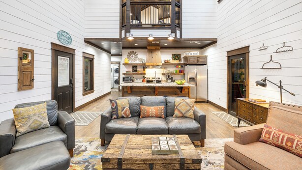 A spacious living area with a central gray leather sofa, a wooden coffee table, and a kitchen in the background.