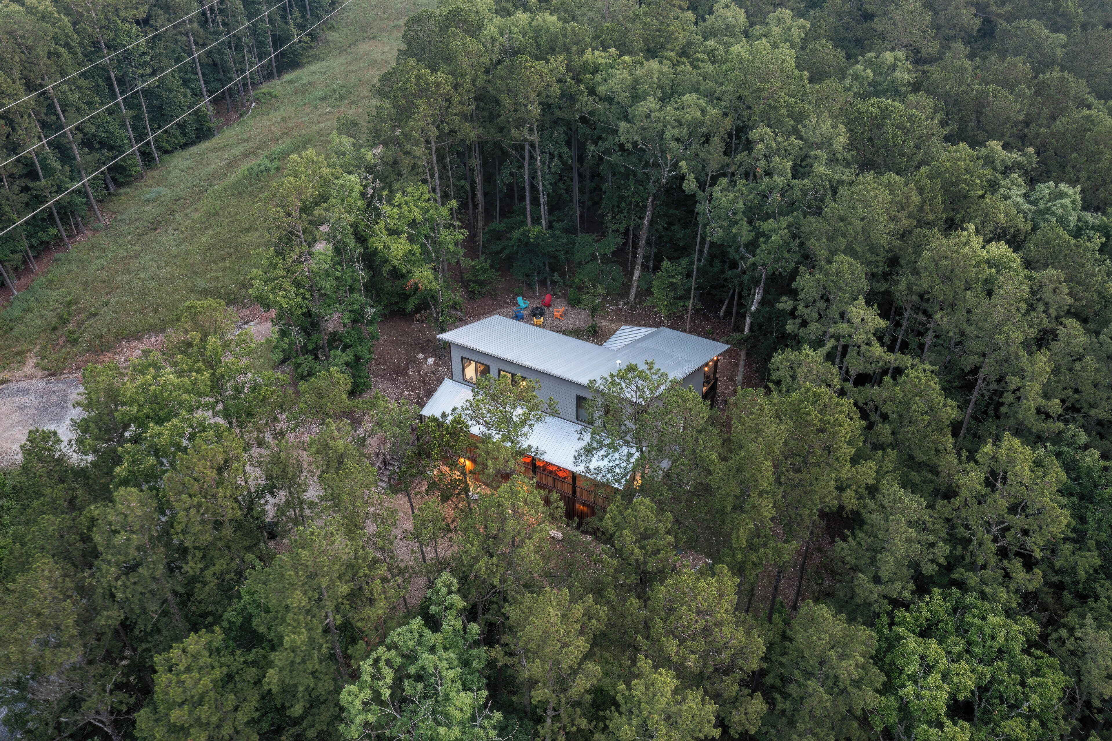 A modern house surrounded by dense forest, with a clear view of the surrounding trees and a visible road to the left.