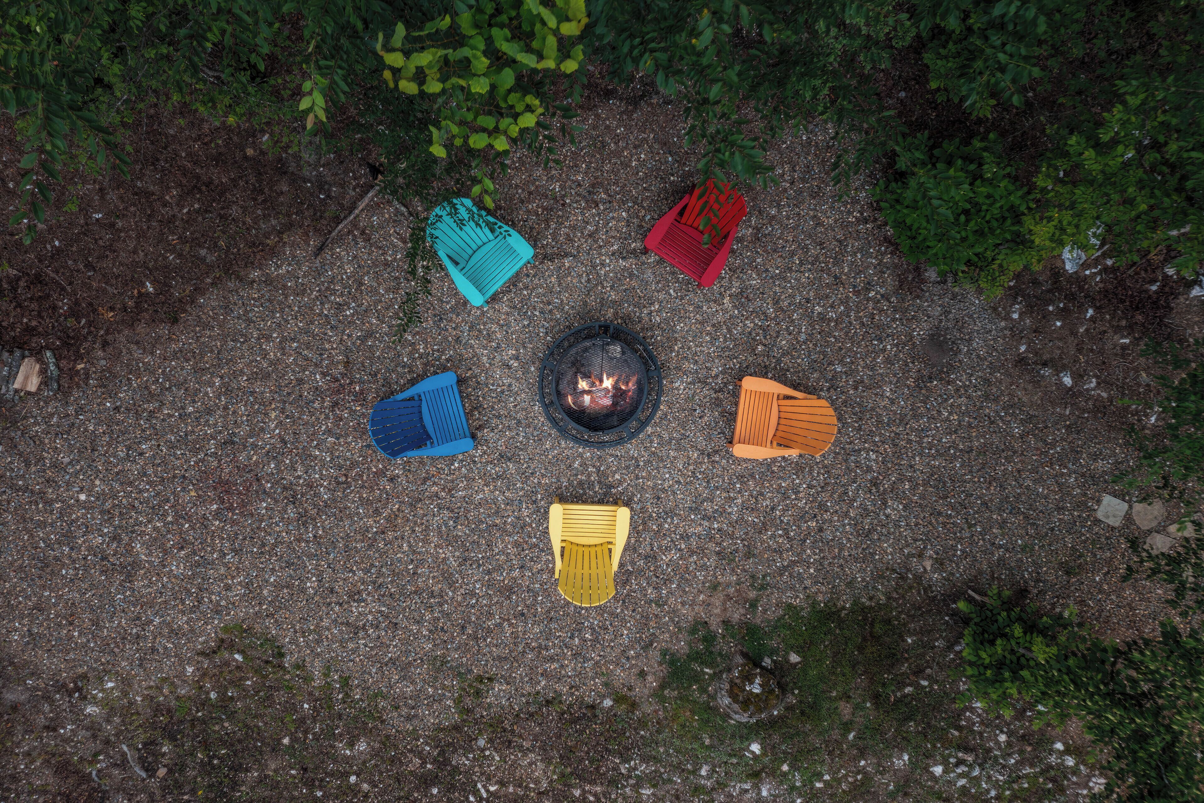An aerial view of a campsite with a fire pit surrounded by five colorful Adirondack chairs.